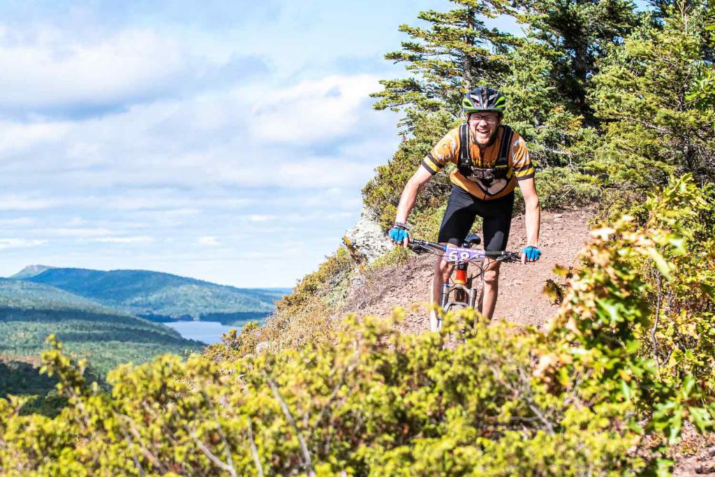 A mountain biker smiles in front of a huge view from a cliff-side trail.